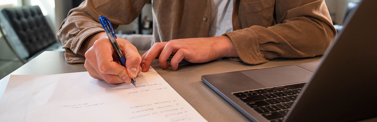 A person sitting at a table with a pen and paper and laptop working on their letter of introduction for a teaching job.